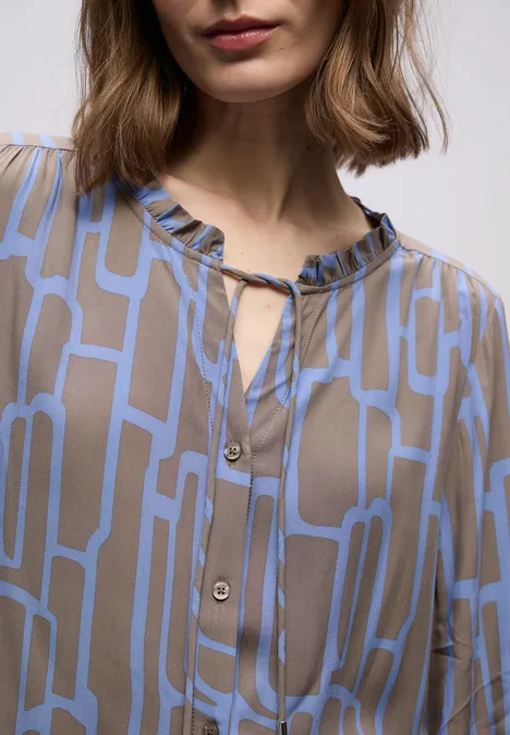 Nahaufnahme einer Frau in einer beigen Bluse mit blauem, geometrischem Muster und Rüschenkragen.